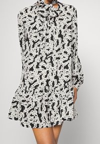 Vestido negro con estampado de cadenas blancas, con un escote en forma de lazo, mangas largas y un dobladillo acampanado con detalles de volantes. Tela suave y ligera.