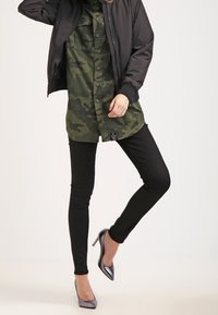 Schwarze Bomberjacke über einem olivgrünen Camouflage-Shirt, kombiniert mit schwarzen Skinny Jeans und grauen High Heels. Glatte Texturen und taillierte Passform.