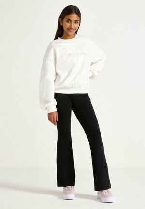Felpa bianca con scritta "Bonjour" colorata, abbinata a pantaloni a zampa nera e sneakers rosa chiaro. Texture liscia, vestibilità rilassata.