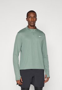 Grünes langärmliges Sportoberteil mit einem Reißverschlusskragen und Daumenlöchern, ausgestattet mit einem kleinen weißen Swoosh-Logo auf der linken Brust.