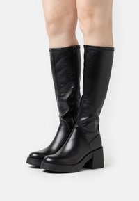 Bottes noires montantes en cuir lisse, avec un talon bloc épais, une fermeture éclair latérale et une texture légèrement plissée sur la tige.