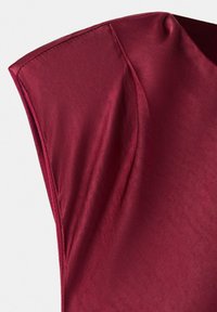 Primo piano di un indumento rosso senza maniche che mostra spalle strutturate e dettagli di cuciture su tessuto liscio.