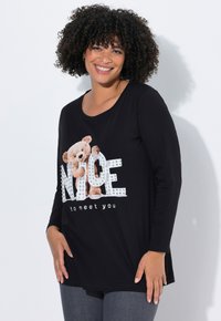 Czarna koszula z długim rękawem z grafiką misia i literami zdobionymi kryształkami układającymi się w słowo „NICE”, poniżej napis „miło cię poznać”.