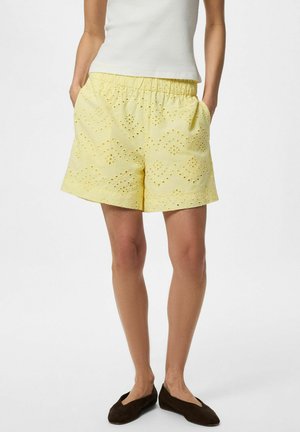 Femme portant un short en tissu œillet jaune clair avec une taille élastique et des poches, associé à des ballerines noires et un haut blanc sans manches.