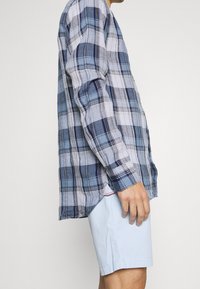 Långärmad rutiga skjorta i blå, vit och marin mönster, tillverkad av lättviktsmaterial. Kombinerad med ljusblå shorts, sidovisning.