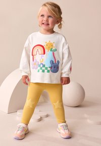 Chemise blanche à manches longues avec motifs brodés colorés, leggings jaunes ornés d'appliques en forme d'étoiles, et baskets pastel avec des accents gris.