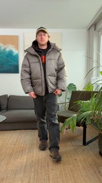 Giacca imbottita grigia sopra una felpa nera con cappuccio, pantaloni grigio scuro e scarpe marroni; ambientazione interna con piante e arte contemporanea sullo sfondo.