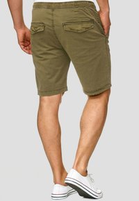 Olivgrüne Baumwollshorts mit elastischem Bund, zwei Gesäßtaschen und lockerem Schnitt. Kombiniert mit weißen Canvas-Schuhen.