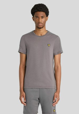 Ung mand med kort krøllet hår iført et matchende gråt t-shirt- og shorts-sæt med en lille gul fuglelogo på begge beklædningsgenstande.
