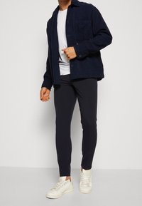 Uomo che indossa un giacca in corduroy blu scuro, una maglietta bianca, pantaloni neri slim e sneaker bianche, in piedi contro uno sfondo chiaro e neutro.
