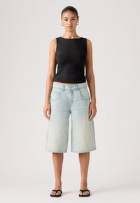 Γυναίκα με αμάνικο μαύρο τοπ, ανοιχτό μπλε τζιν σόρτς μέχρι το γόνατο, μαύρα σανδάλια και χρυσούς κρίκους στα αυτιά, στέκεται μπροστά σε απλό φόντο.