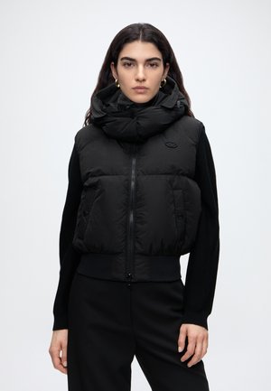 Donna con capelli scuri indossa un gilet imbottito nero con collo alto sopra una maglia nera a maniche lunghe, in piedi davanti a uno sfondo chiaro e uniforme.