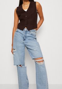 Brun ærmeløs vest med fem knapper, kombineret med lyseblå distressed jeans med store revne knædetaljer.