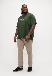 T-shirt en coton vert foncé avec un texte graphique blanc, associé à un pantalon beige et des baskets noires avec des accents marron.