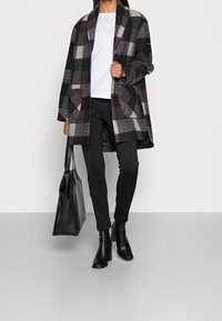 Karierten Wollmantel in Schwarz, Grau und Creme, mit übergroßen Taschen. Kombiniert mit schwarzen Slim Pants, Stiefeletten und einer schwarzen Ledertasche.