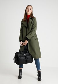 Olive green trench coat with a wide collar, black quilted handbag, blue jeans, and black ankle boots. Simple, functional design.