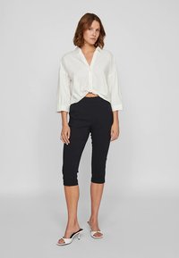 Valge pikkade varrukatega nööpidega särgikleit, mis on vöökohast sõlmitud, koos kohandatud mustade lühendatud pükstega ja avatud varbaga valgete kontsakingadega.