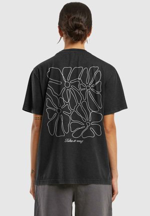 Person with hair in bun wears black t-shirt featuring white abstract floral design and "Take it easy" text on the back.