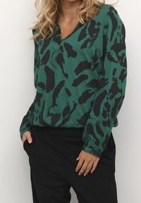 Blouse verte avec des motifs abstraits noirs, décolleté en V, manches longues et coupe décontractée. Le tissu a une texture douce. Associée à un pantalon noir.
