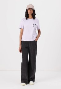 Lilac short-sleeve t-shirt with a front pocket graphic, paired with black wide-leg trousers. The outfit is completed with a beige baseball cap.