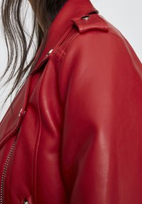 Gros plan sur l'épaule et le cou d'une femme portant une veste en cuir rouge avec des boutons-pression argentés et une fermeture éclair.