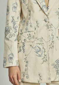 Blazer in lino beige con ricami floreali blu e verdi. Presenta una chiusura a singolo bottone e un colletto con rever. Tessuto testurizzato.