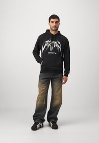 Felpa nera con grafica frontale, abbinata a jeans in denim chiaro a scuro con effetto usurato e sneakers nere con dettagli bianchi.