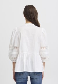 Blusa bianca con maniche a sbuffo, dettagli in pizzo, orlo plissettato, tessuto morbido e vestibilità comoda, indossata con jeans blu. Viene mostrata la vista posteriore.