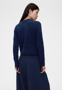 Tamsiai mėlynas vilnos mišinys megztinis su trumpu kirpimu, apvali iškirptė ir siauri rankogaliai. Tekstūruota medžiaga, dėvima su aukšto liemens dryžuotomis kelnėmis.