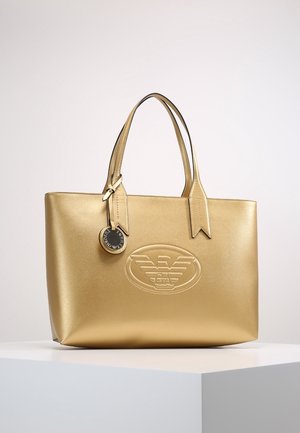 Gouden leren tote-tas met twee schouderriemen, geperst logo aan de voorkant en een ronde metalen hanger aan één riem, tentoongesteld op een wit voetstuk.