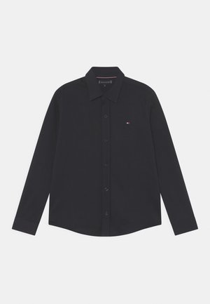 Chemise noire manches longues boutonnée avec col, petit logo brodé rouge, blanc et bleu sur la poitrine gauche, posée à plat sur fond blanc.