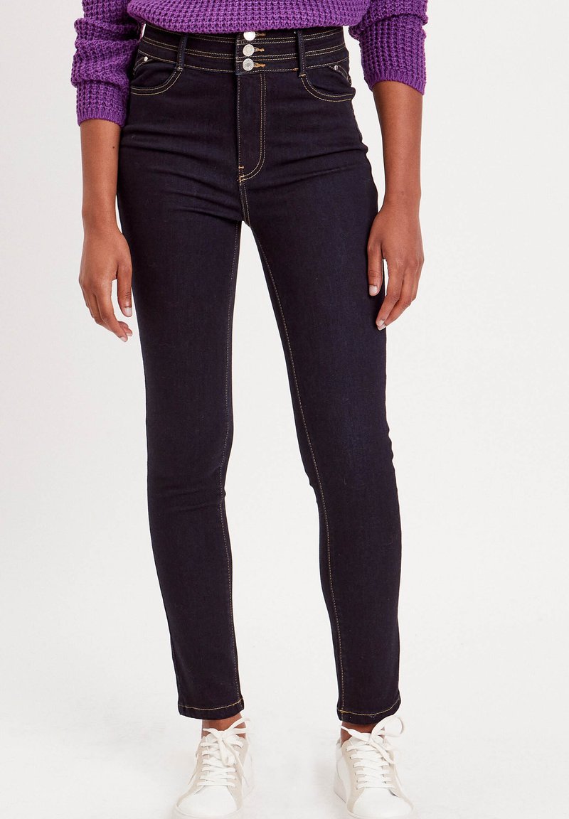 Jeans en denim foncé taille haute avec une texture lisse, cinq poches et trois boutons argentés à l'avant ; associés à des baskets blanches.