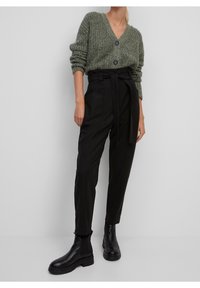 Cardigan vert en tricot à col en V avec deux boutons noirs, associé à un pantalon noir taille haute avec une ceinture à nouer et des bottines noires plates.