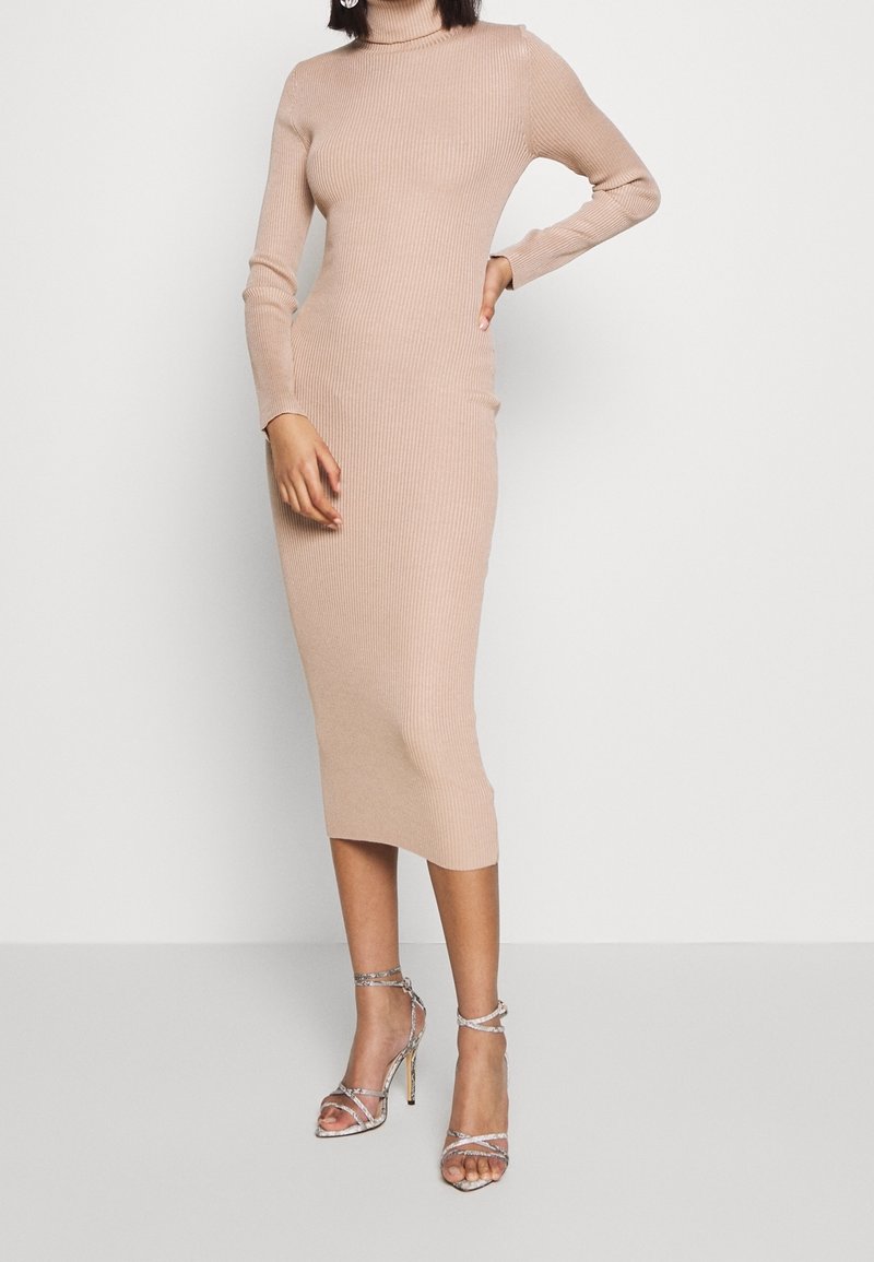 Abito a maniche lunghe in maglia a coste di colore beige, con una silhouette aderente che arriva sotto il ginocchio, abbinato a sandali alti argentati con strappi.