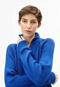 Blauer gerippter Strickpullover mit hohem Kragen und Reißverschluss. Der Stoff wirkt strukturiert und weich und weist markante Nahtdetails auf.