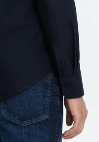 Tmavě námořnická košile s dlouhým rukávem, s knoflíkovým manžetem a lehce zakřiveným lemem, doplněná modrými džínami s kapsou na zadní straně.