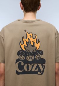 Béžové bavlněné tričko s grafickým designem táboráku a oranžovými plameny nad slovem "Cozy" v tučném černém písmu.