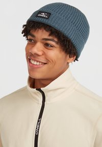 Gorra de punto azul con textura acanalada y un parche negro con el logo de O'Neill en la parte frontal, llevada con una chaqueta de forro polar beige con cremallera.