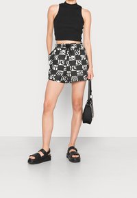 Personne portant un crop top noir sans manches, un short à motifs noir et blanc, des sandales noires, et tenant un sac à main noir avec une bandoulière chaîne.