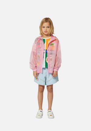 Veste à capuche translucide rose sur une chemise graphique multicolore, associée à un short bleu clair avec des patchs décoratifs et des chaussures blanches.