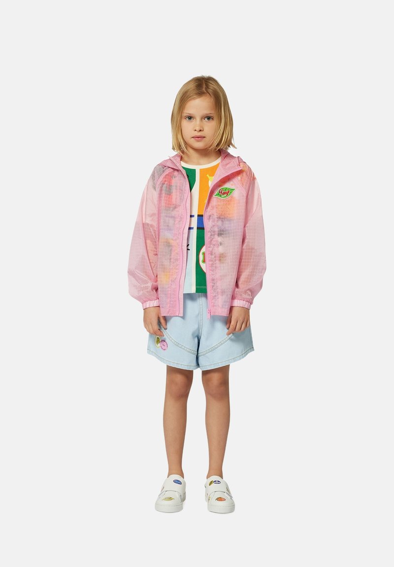 Roze doorschijnende hoodie-jack over een multi-kleurig graphic shirt, gecombineerd met lichtblauwe shorts met decoratieve patches en witte schoenen.