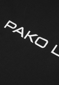 Czarna tkanina z wyraźnym białym haftem słowa "PAKO" w nowoczesnej czcionce bezszeryfowej, prezentująca gładką fakturę i czyste linie.