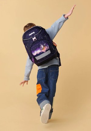 Child wearing dark starry backpack with fairy design, posing with arms outstretched against plain beige background.