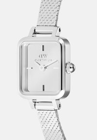 Montre rectangulaire en argent avec bracelet en maille, cadran épuré avec des marqueurs des heures et une couronne pour régler l'heure. Nom de la marque gravé sur le cadran.