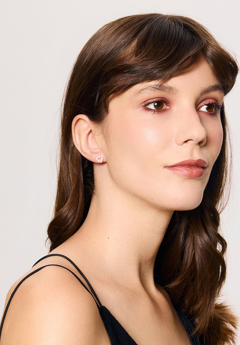 Srebrne kolczyki w kształcie serca, zaprezentowane na modelce, podkreślające wypolerowane wykończenie i ażurowy wzór. Modelka z długimi, ciemnymi włosami.