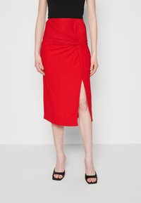 Jupe rouge mi-longue en tissu texturé, avec un détail torsadé à la taille et une fente sur le côté, accompagnée de talons ouverts noirs.