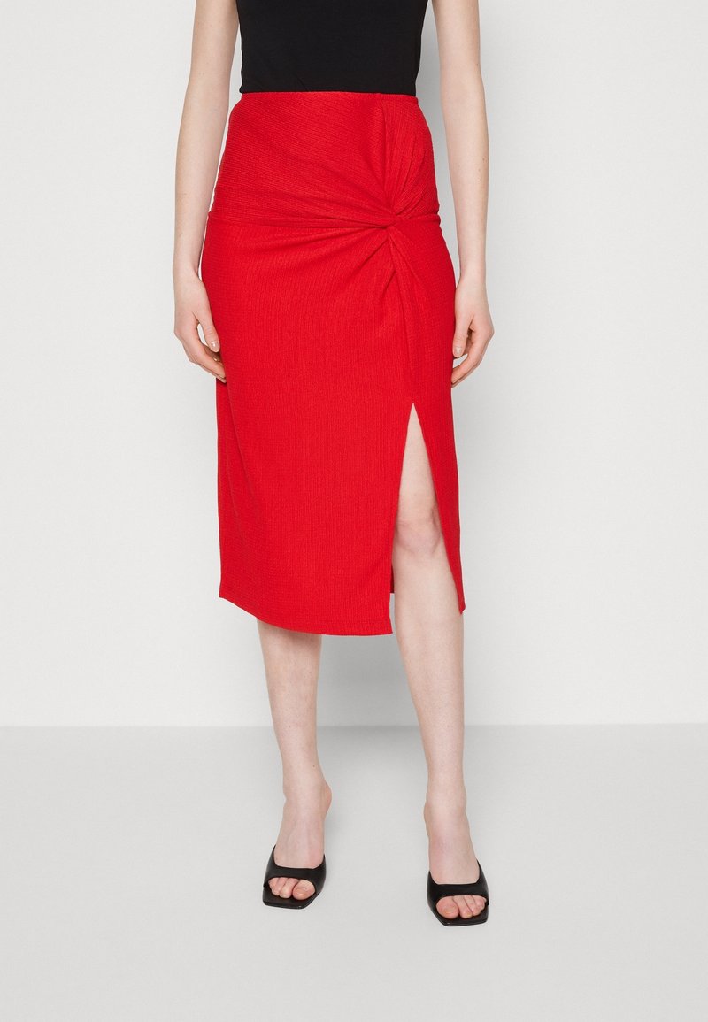 Jupe rouge mi-longue en tissu texturé, avec un détail torsadé à la taille et une fente sur le côté, accompagnée de talons ouverts noirs.