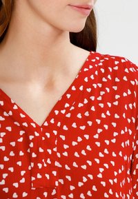 Blouse rouge avec un col en V en tissu texturé. Décorée de petits motifs de cœurs blancs. Matériau doux et léger.