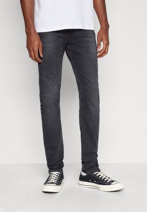 Homme portant un jean slim noir, des chaussettes blanches, des baskets noires basses et un t-shirt blanc, debout devant un fond uni.