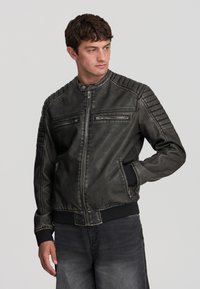Giacca bomber in pelle nera con spalle a coste, tasche sul petto con zip e vita aderente. La texture scucita aggiunge profondità.